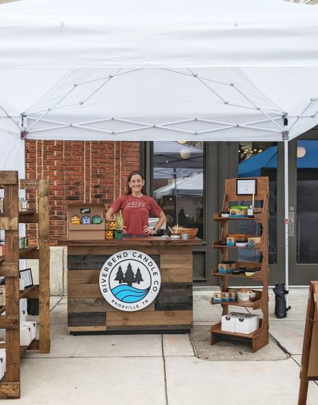 A woman wearing a read shirt stands proudly with her hands on hips behind a wood-clad counter under a white canopy tent. She looks directly into the camera and smiles. Flanking her are rustic wooden shelves that display candles.