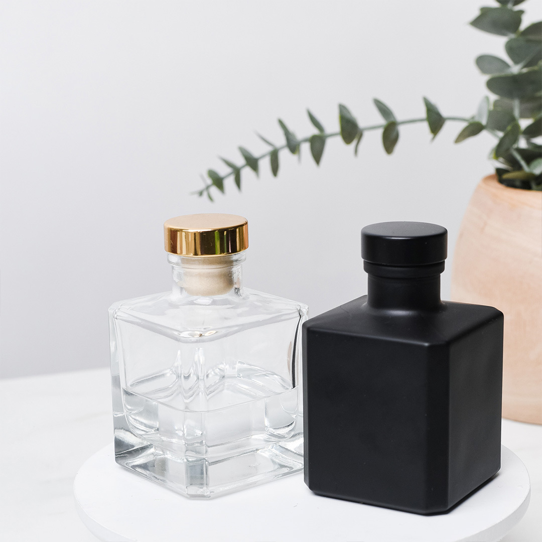 A matte black cube and clear cube reed diffuser bottle on a marble table with eucalyptus in the background.