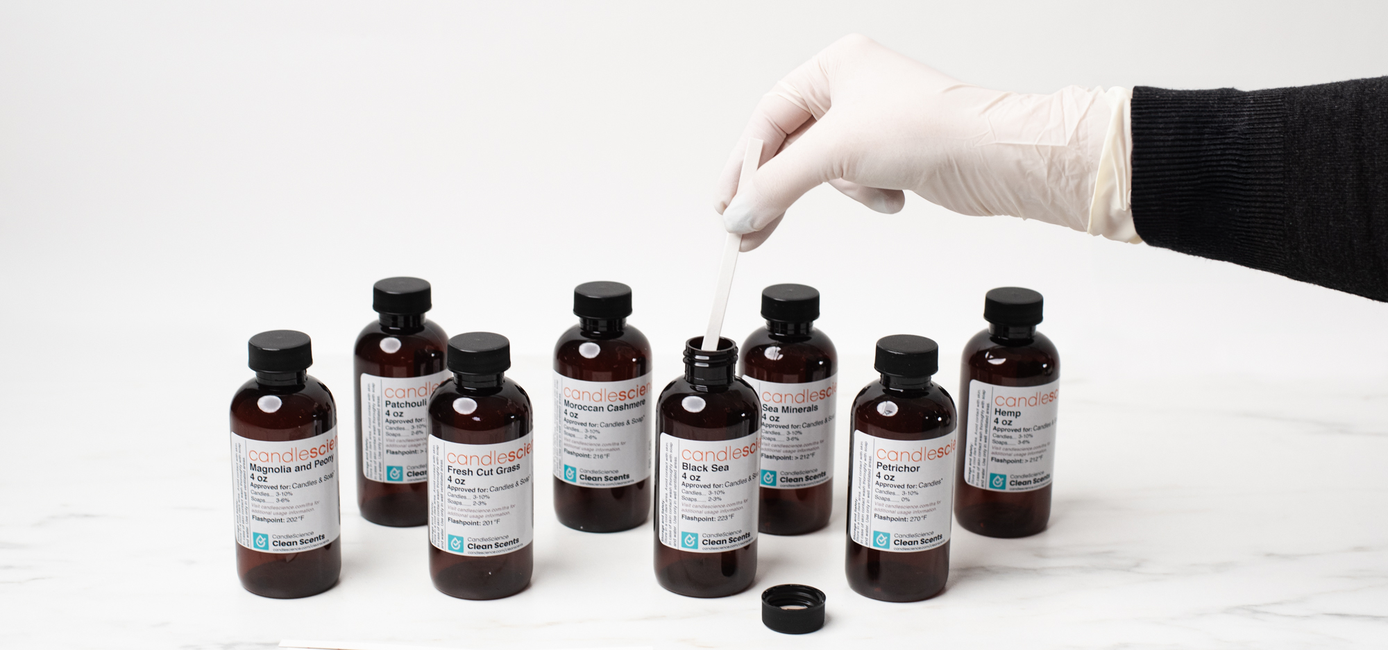 A gloved hand dips a fragrance blotter strip into a bottle of CandleScience fragrance oil