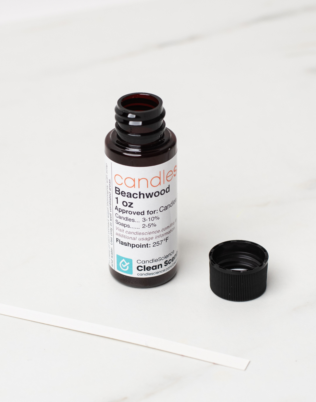 A 1 oz bottle of CandleScience fragrance oil with its lid off and a fragrance blotter strip next to it