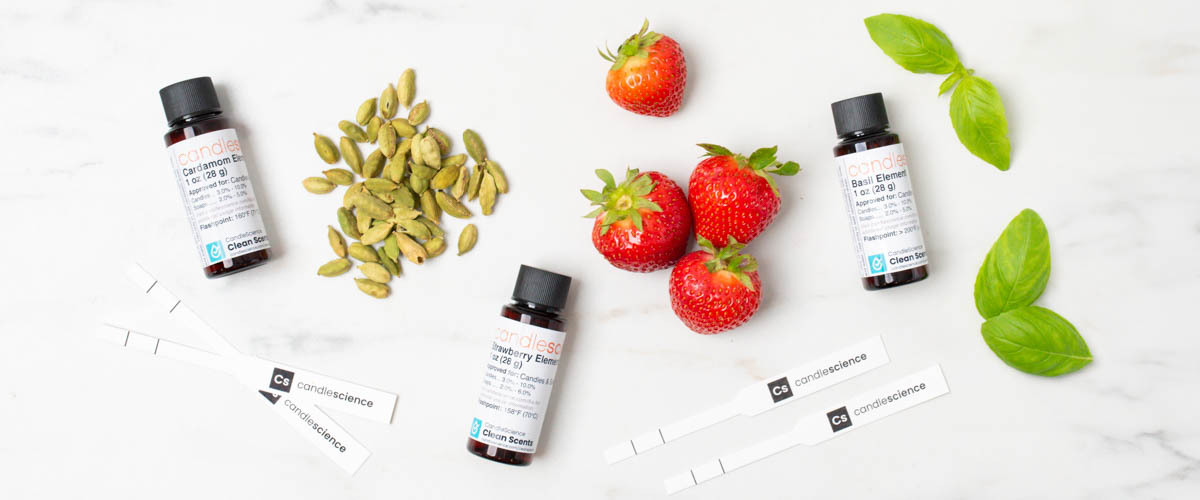 Green cardamom, whole strawberries, and basil leaves on a white marble table with fragrance blotter strips and three 1 oz bottles of fragrance oil.