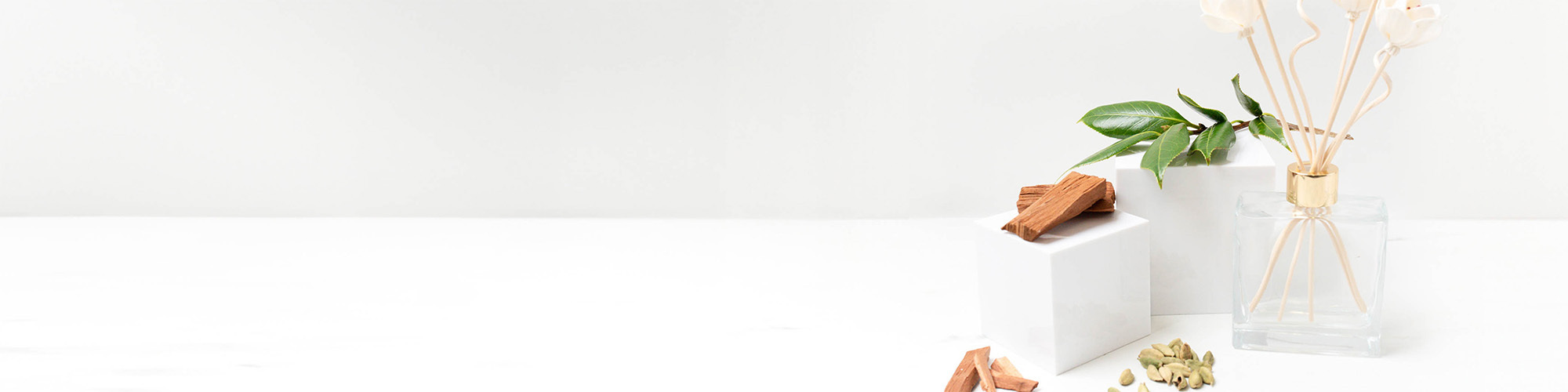 green leaves, sandalwood chips, and cardamom on white marble pillars beside a reed diffuser bottle with spiral and flower reed sticks.